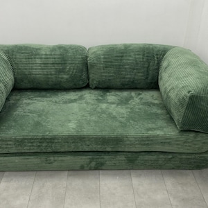 May include: A green corduroy sofa with two back cushions and two armrests. The sofa is a muted green color and has a textured, ribbed appearance. The sofa is placed against a white wall.
