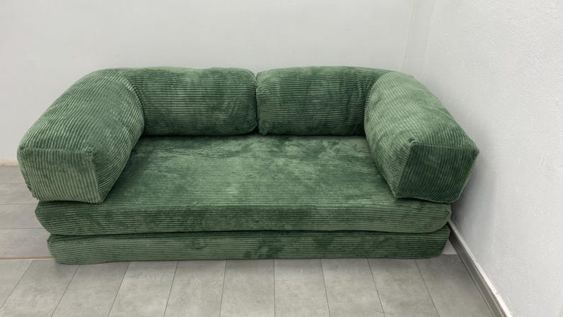 May include: A green corduroy sofa with two back cushions and two armrests. The sofa is a muted green color and has a textured, ribbed appearance. The sofa is placed against a white wall.