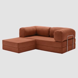 Puede incluir: Un sof&aacute; seccional modular de pana color &oacute;xido con una chaise longue. El sof&aacute; presenta un perfil bajo, reposabrazos redondeados y una estructura met&aacute;lica. La tela tiene una textura acanalada. La chaise longue est&aacute; en el lado izquierdo del sof&aacute;.