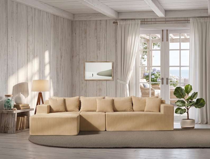 May include: A light beige corduroy sectional sofa with matching throw pillows. The sofa is in a bright, airy living room with white curtains, a wooden side table, and a potted plant. A framed picture hangs on the wall.
