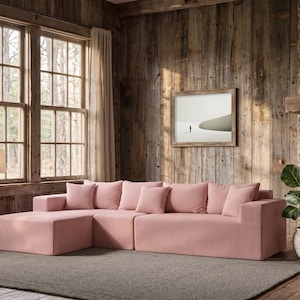 May include: A large, sectional sofa in a soft pink corduroy fabric. The sofa has a chaise lounge and several matching throw pillows. The room features a rustic wooden wall, a framed picture, and a potted plant.