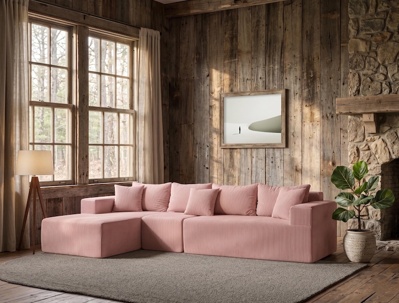 May include: A large, sectional sofa in a soft pink corduroy fabric. The sofa has a chaise lounge and several matching throw pillows. The room features a rustic wooden wall, a framed picture, and a potted plant.
