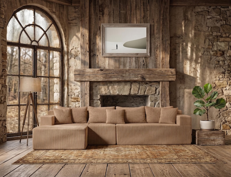 May include: A modern, modular sectional sofa in a neutral beige corduroy fabric. The sofa is arranged in an L-shape and includes several matching throw pillows. The room features a rustic aesthetic with a large window, wooden floors, and a stone fireplace.