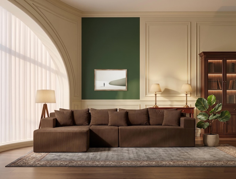 May include: A brown corduroy sectional sofa with multiple cushions. The sofa is placed on a patterned rug in a room with a green accent wall and a framed picture. A wooden bookcase and a potted plant are also visible.