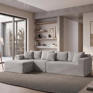 May include: A modern, light gray sectional sofa with multiple plush cushions. The sofa features a corduroy texture and is placed on a gray rug. A wooden tripod floor lamp and a potted plant are also visible in the room, creating a cozy living space.