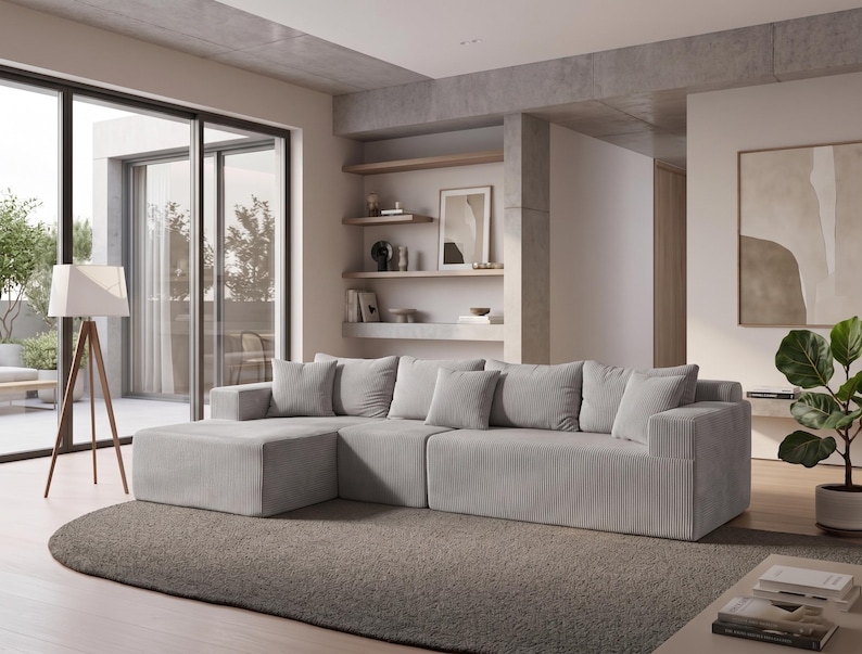 May include: A modern, light gray sectional sofa with multiple plush cushions. The sofa features a corduroy texture and is placed on a gray rug. A wooden tripod floor lamp and a potted plant are also visible in the room, creating a cozy living space.