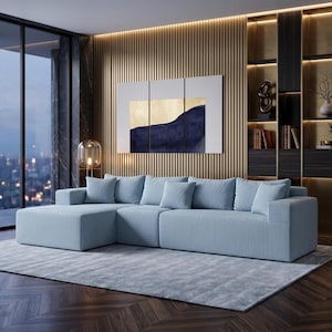 May include: A light blue sectional sofa with matching throw pillows sits on a light gray rug. The room features a large window, a wooden accent wall, and a modern painting with gold and navy blue tones. A decorative lamp and a potted plant add to the decor.