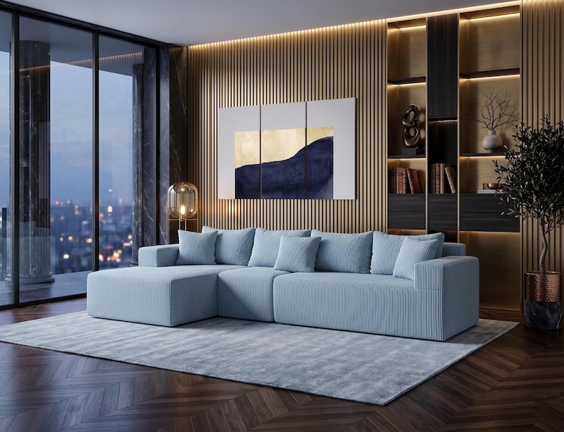 May include: A light blue sectional sofa with matching throw pillows sits on a light gray rug. The room features a large window, a wooden accent wall, and a modern painting with gold and navy blue tones. A decorative lamp and a potted plant add to the decor.