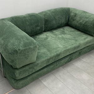May include: A green corduroy sofa with a rounded back and armrests. The sofa has a low profile and a simple, modern design. The frame is a silver metal. The cushions are a solid green color.