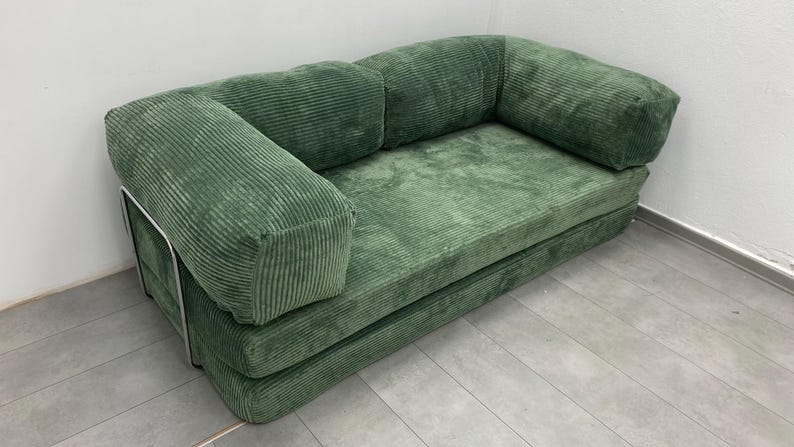 May include: A green corduroy sofa with a rounded back and armrests. The sofa has a low profile and a simple, modern design. The frame is a silver metal. The cushions are a solid green color.