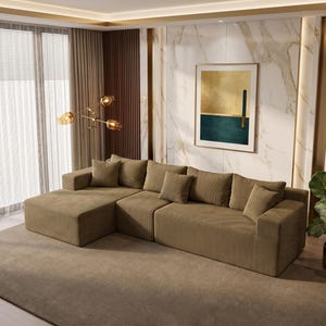 May include: A large, olive-green sectional sofa with multiple throw pillows sits on a matching area rug. The sofa has a corduroy texture. A framed abstract art piece hangs on the wall, and a potted plant is to the right.