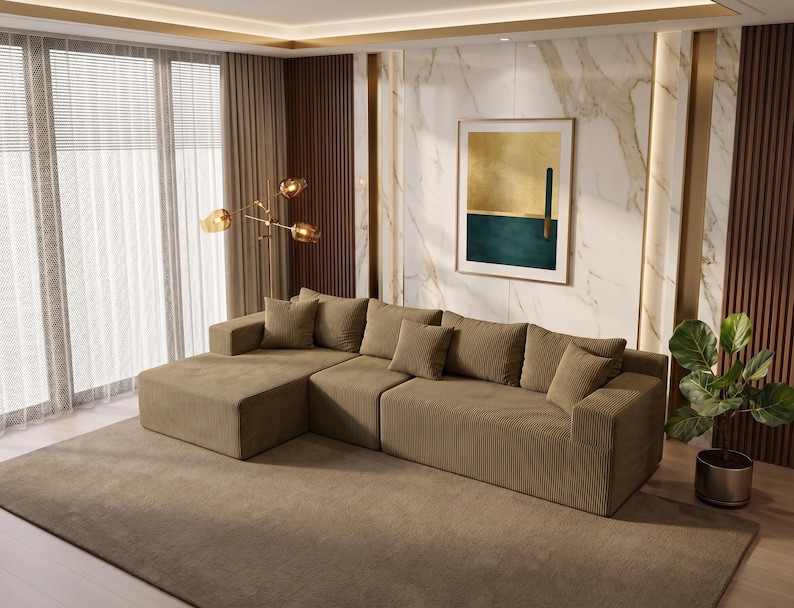 May include: A large, olive-green sectional sofa with multiple throw pillows sits on a matching area rug. The sofa has a corduroy texture. A framed abstract art piece hangs on the wall, and a potted plant is to the right.