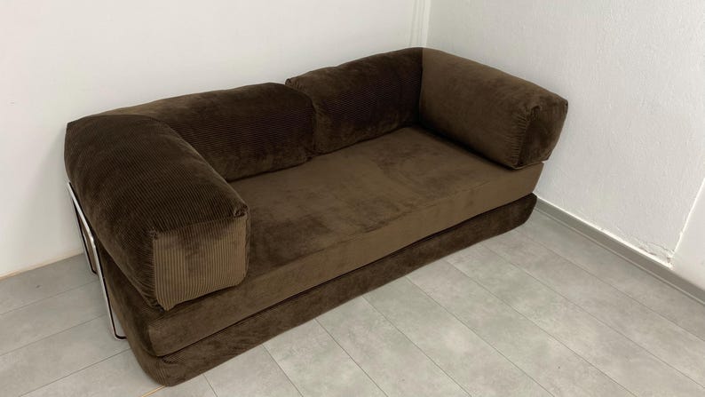 May include: A brown corduroy sofa with a low profile and rounded armrests. The sofa has a rectangular seat and back cushions. The frame appears to be metal. The sofa is placed against a white wall.