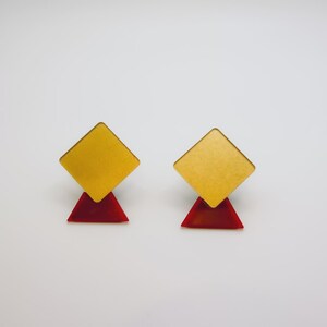 Pode incluir: Um par de brincos de tarracha com uma peça superior quadrada em tom dourado fixada a uma base triangular menor em vermelho translúcido. O design geométrico combina um acabamento metálico no quadrado com um material vermelho semitransparente.