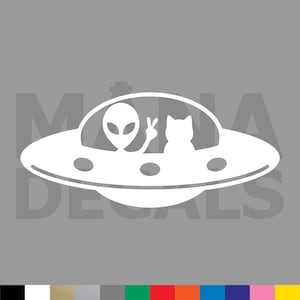 May include: A white vinyl decal featuring a flying saucer with an alien making a peace sign and a cat sitting inside the cockpit. The design includes circular details along the edge of the saucer. The text MANIA DECALS is visible in the background.