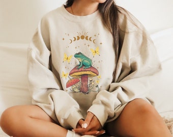 Puede incluir: Una sudadera beige con un estampado gráfico de una rana verde sentada en un hongo con mariposas amarillas. El gráfico también incluye un diseño de sol y luna con estrellas.
