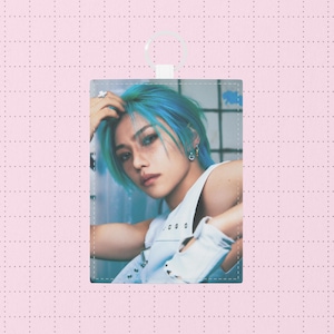 Könnte beinhalten: Eine rechteckige Karte mit einer Person mit blauen Haaren und einem weißen ärmellosen Oberteil. Die Karte hat oben eine weiße Schlaufe und befindet sich vor einem rosa karierten Hintergrund.