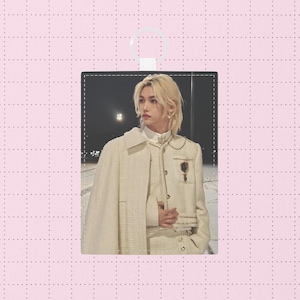 Könnte beinhalten: Ein rechteckiger cremefarbener Anhänger mit einem Foto einer Person in einem cremefarbenen Anzug mit Umhang. Die Person hat blondes Haar und schaut zur Seite. Der Anhänger hat oben eine weiße Schlaufe zum Aufhängen.