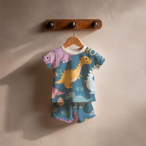 May include: A two-piece children's pajama set featuring a teal background with colorful dinosaur illustrations. The short-sleeved top and shorts are hanging on a wooden hanger. The top has a white collar and the word "ROAR" is visible.