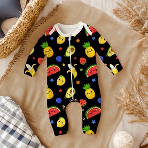 May include: Black baby onesie with a white zipper and trim, featuring a colorful pattern of smiling fruits including pineapples, lemons, avocados, strawberries, bananas, and watermelon. The design also includes blue stars and blueberries.