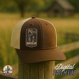 Op de afbeelding: Bruine en beige trucker cap met een bruin patch waarop "Bayed Up" staat en een cartoon wasbeer.