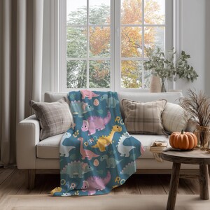 May include: A blue throw blanket with a repeating pattern of cartoon dinosaurs in pink, yellow, white, and green. The word "ROAR" and "DINO" are also printed on the blanket. The blanket is draped over a white couch.