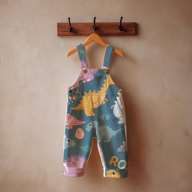 May include: Blue and white baby overalls with a dinosaur print. The overalls feature a variety of colorful dinosaurs, eggs, and the word "RO" on the leg. The overalls are hanging on a wooden hanger.