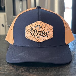 May include: Navy blue and tan baseball cap with a curved brim. The cap features a tan mesh back and a navy blue front panel. A tan leather patch on the front displays the words "Master Baiter" with a fish graphic.