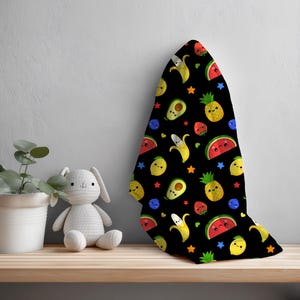 May include: Black baby blanket with a repeating pattern of smiling cartoon fruits including bananas, avocados, pineapples, strawberries, lemons, and watermelon. The blanket also features blue stars and blueberries.