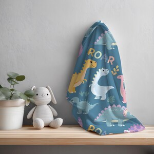 May include: A blue blanket with a dinosaur pattern and the word "ROAR" in yellow. The blanket features various cartoon dinosaurs in yellow, white, and pink. A stuffed bunny and a potted plant are on a wooden shelf.