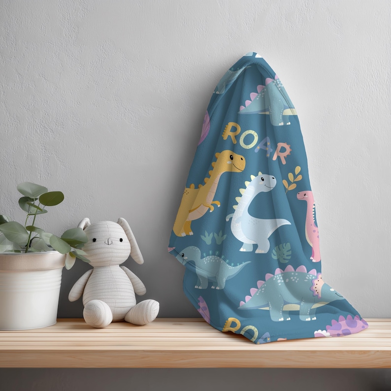 May include: A blue blanket with a dinosaur pattern and the word "ROAR" in yellow. The blanket features various cartoon dinosaurs in yellow, white, and pink. A stuffed bunny and a potted plant are on a wooden shelf.