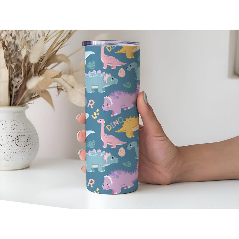 May include: A stainless steel tumbler with a teal background and a repeating pattern of colorful dinosaurs, eggs, and the word "DINO". The tumbler is cylindrical and has a silver lid.