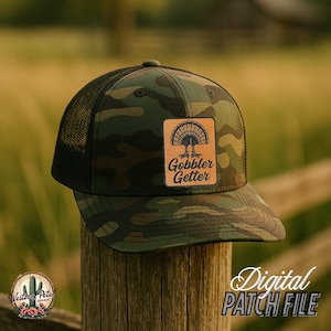 Puede incluir: Una gorra de camionero de camuflaje con un parche de cuero marrón que dice "Gobbler Getter" y presenta una silueta de pavo.