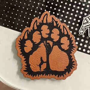 May include: A brown leather paw print with a black silhouette of a tree and a dog inside. The paw print is a decorative item.