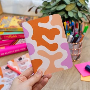 May include: A small notebook with a colourful abstract design featuring orange, pink and white shapes. The notebook is being held in a hand.