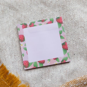 May include: A square notepad with a pink border featuring red strawberries, green leaves, and white flowers. The centre of the notepad is a blank, light purple space for writing. The notepad is on a textured, beige surface.