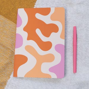 May include: A white notebook with a cover featuring an abstract pattern of orange, peach, and pink shapes. A pink pen is lying next to the notebook.