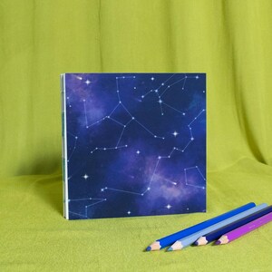 May include: A square notebook with a dark blue cover featuring a constellation design. Three coloured pencils in blue, purple, and violet are arranged on a green surface next to the notebook.