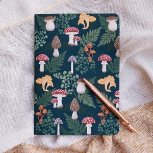 May include: A dark blue notebook with a mushroom and fern pattern. The notebook is closed and has a copper-colored pen lying on top of it.