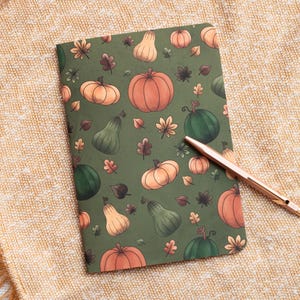 May include: A green journal with a autumn-themed design featuring pumpkins, gourds, and leaves. The journal is lying on a beige blanket with a gold pen next to it.