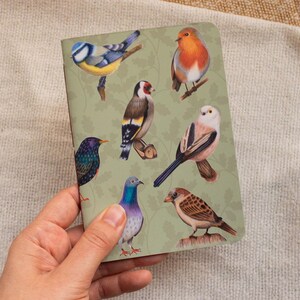 Op de afbeelding: Een rechthoekig notitieboek met een lichtgroene kaft voorzien van een herhalend bladpatroon en gedetailleerde illustraties van diverse vogels, waaronder een pimpelmees, een roodborstje, een putter, een staartmees, een spreeuw, een duif en een mus.