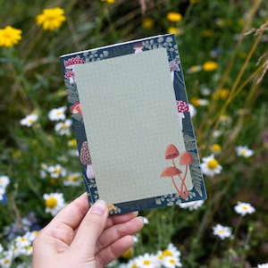 Könnte beinhalten: Ein kleines, blaues Notizbuch mit einer grünen, gepunkteten Seite und einem Pilzrand. Der Rand zeigt rote und weiße Pilze mit braunen Stielen.