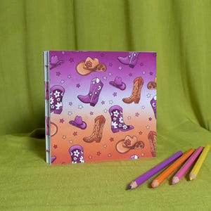 May include: A square notebook featuring a pattern of cowboy boots, hats, and stars in purple, orange, and brown. The notebook is on a green surface with four coloured pencils.