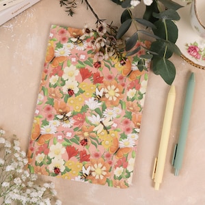 以下が含まれることがあります： ピンクの背景に鮮やかな花柄が描かれた長方形のノート。オレンジ色の蝶、マルハナバチ、赤いテントウムシのイラストが装飾されています。ノートの横には、淡い黄色のペンとセージグリーンのペンが置かれています。