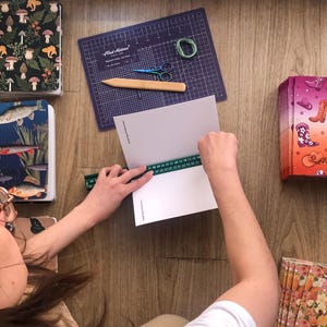 May include: A person uses a green ruler to measure a white paper booklet. Nearby are stacks of notebooks with mushroom, fish, cowboy boot, and floral patterns, a purple cutting mat, a wooden bone folder, small scissors, and green thread.