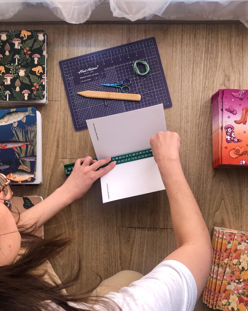 May include: A person uses a green ruler to measure a white paper booklet. Nearby are stacks of notebooks with mushroom, fish, cowboy boot, and floral patterns, a purple cutting mat, a wooden bone folder, small scissors, and green thread.