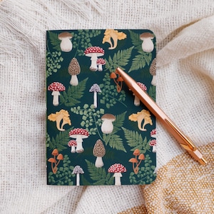 May include: A dark green notebook with a mushroom and fern pattern. The notebook is closed and has a gold pen on top of it.