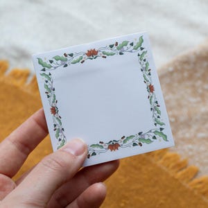 May include: Square white sticky note with a decorative border of green leaves, red flowers, and purple accents. The floral design frames the blank space, ideal for jotting down notes or reminders.