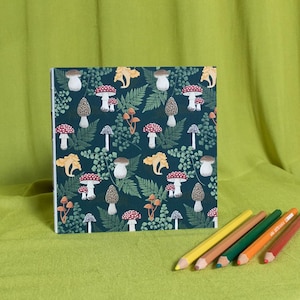 May include: A notebook with a dark teal mushroom pattern, featuring red, white, and brown mushrooms, and yellow ferns. Six coloured pencils in yellow, brown, green, orange, and red are in the foreground.