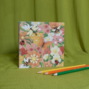 May include: A square notebook with a floral design featuring butterflies, bees, and ladybirds. The notebook is accompanied by three coloured pencils: yellow, orange, and green. The background is a solid green fabric.